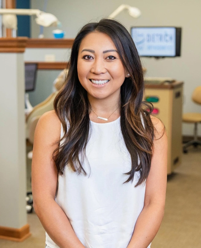 Professional portrait of smiling woman in white tank top, orthodontic office setting with shelving and monitor background.